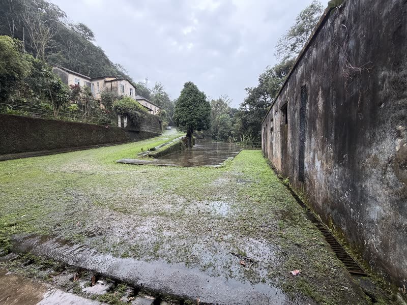 Pátio gramado entre dois muros de alvenaria, mata e morro ao fundo